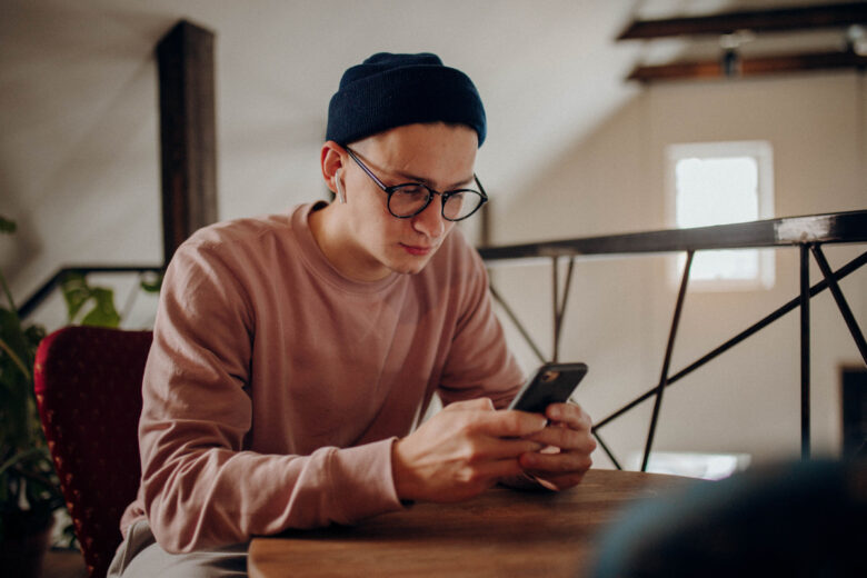Escolha de um celular moderno: o que levar em conta? Jovem de visual casual e óculos usando smartphone em um ambiente interno moderno.